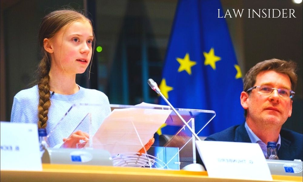 Greta Thunberg alongside a man giving a speech at a conference, wearing a blue Top, a blue colored flag in the background