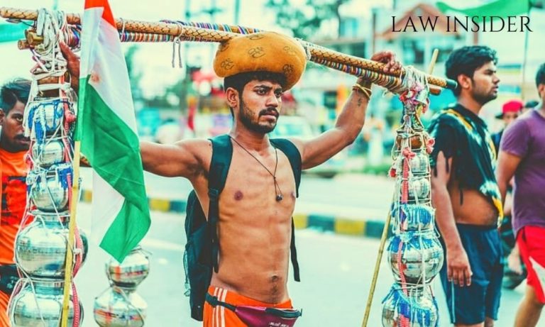 A boy on Kanwar yatra, shirtless in day light wearing a turbun, carrying a black bag, has a wooden stick on his head with steel pots along with them